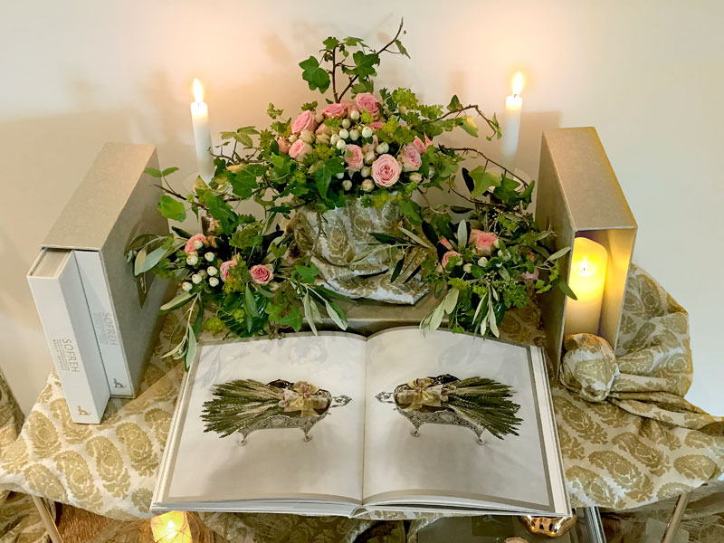 several volumes of the book with softly illuminated delicate bouquets of pink spray roses, hypericum and foliage, and lengths of elegantly draped fine textiles