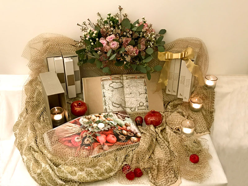 several volumes of the book with a bouquet of Silver Lace roses and foliage, softly draped gold mesh and sculptured pomegranates